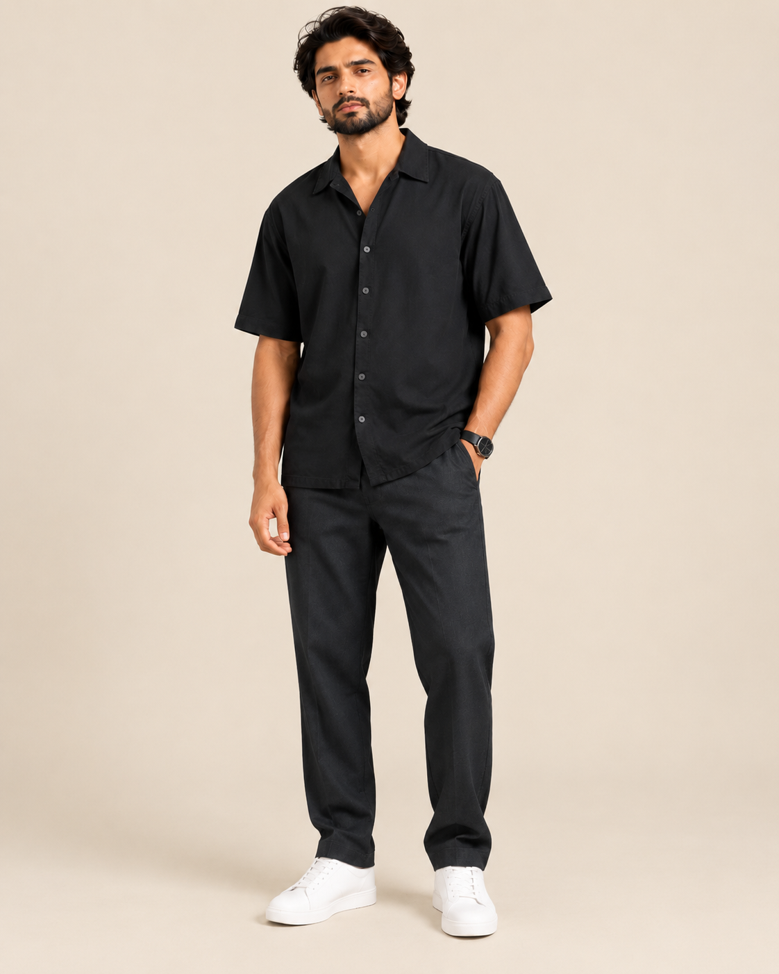 A man modeling a black short-sleeve button-up shirt and matching black trousers, paired with white sneakers, against a solid beige studio background.